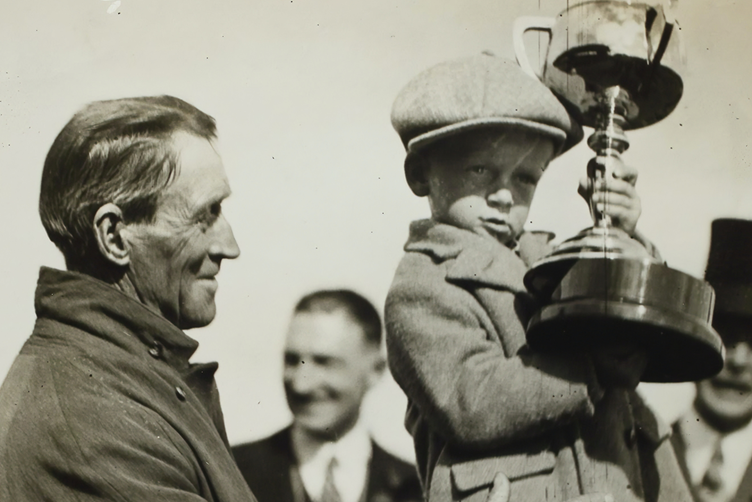 An old photograph of a man holding a boy as he holds onto a large trophy